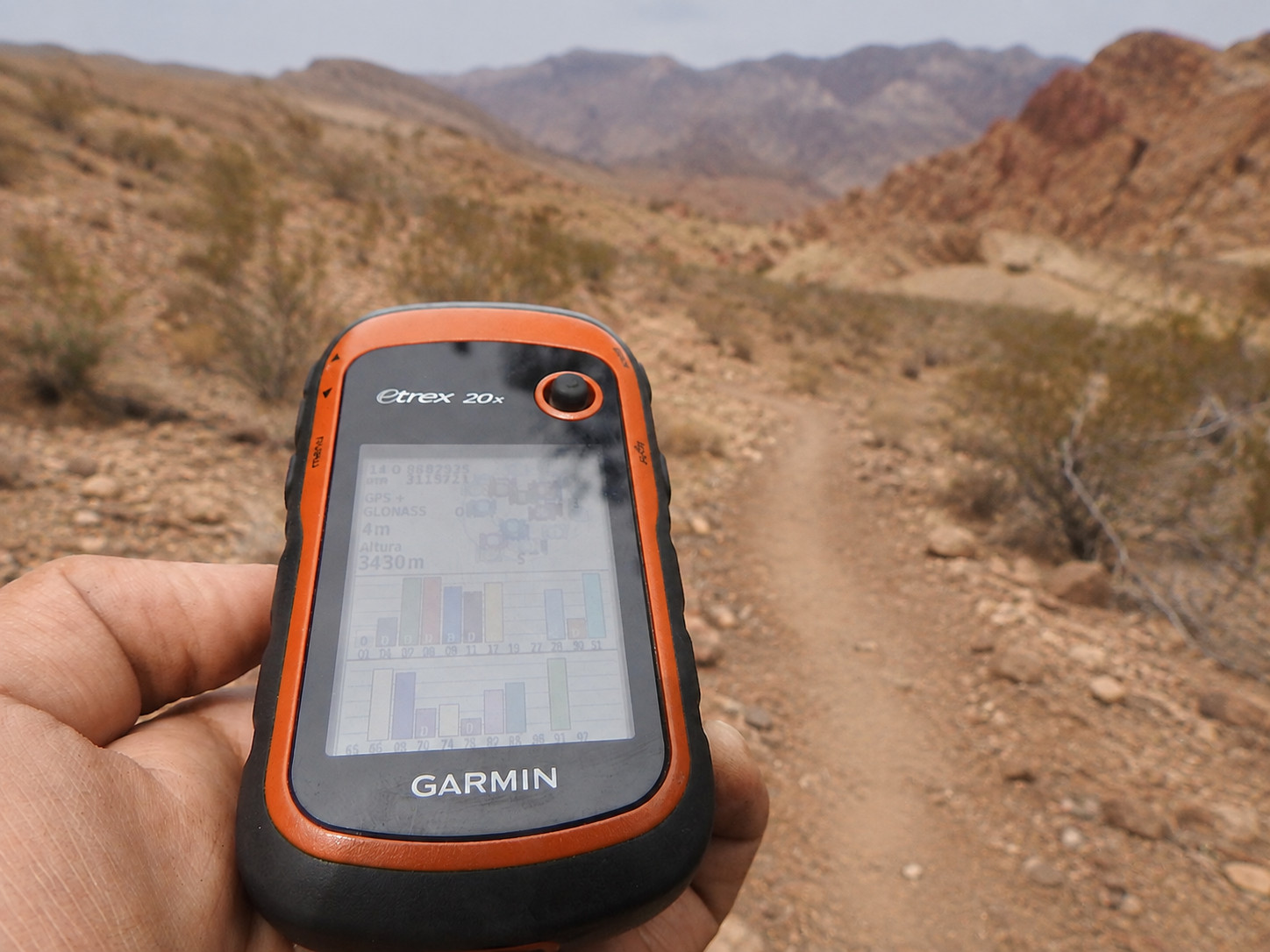 Hand holding a Garmin eTrex GPS unit showing coordinates on a rocky desert trail with red rock mountains behind